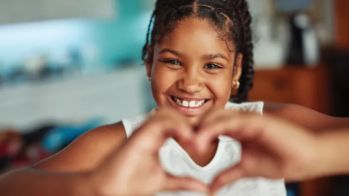 A girl with medium-dark skin tone smiles and makes a heart shape with her hands. 