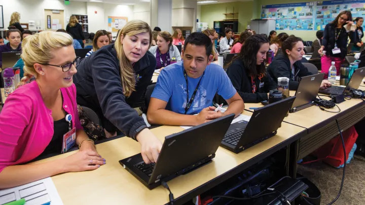 In a classroom, a light skin toned woman points to a computer screen. A light skin toned woman and a medium skin toned man look on.