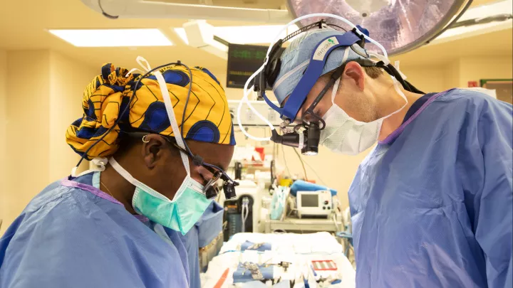 Two masked surgeons wearing full PPE and magnifying eyewear look down in surgical operating room in hospital