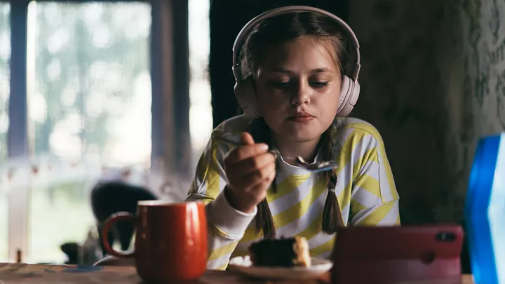Girl in headphones eats a piece of cake while looking down at her mobile phone