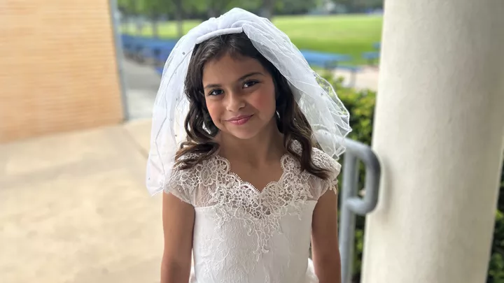 A young girl with medium skin tone wearing a white lace dress and veil smiles at the camera with a grass lawn in the background