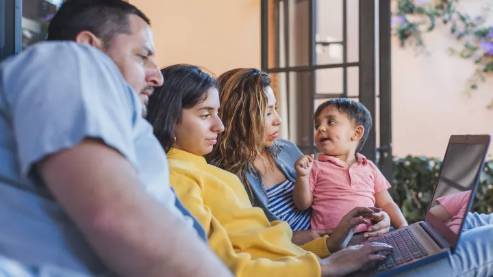 Medium skin-toned family of four sits on couch while using laptop
