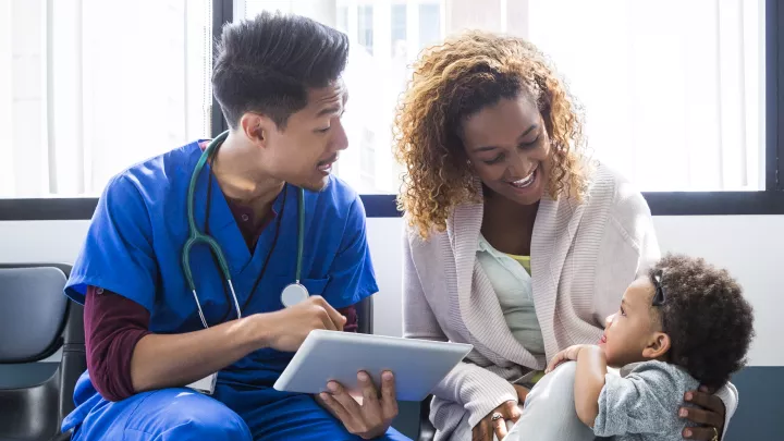 xtA medium skin toned man in blue scrubs and a stethoscope holds a digital tablet and talks to a medium-dark skinned toddler and her dark skin toned mother.