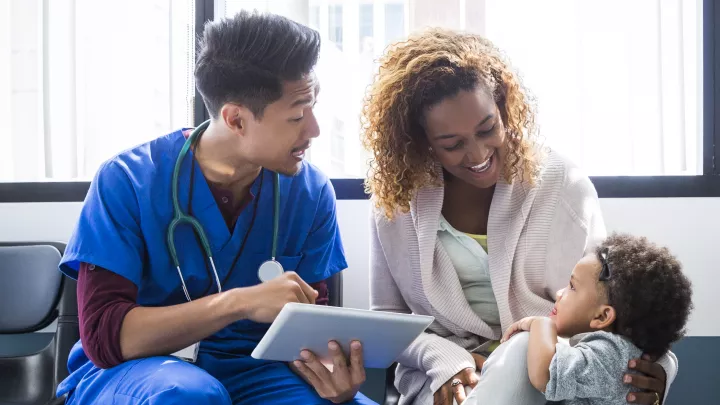 A medium skin toned man in blue scrubs and a stethoscope holds a digital tablet and talks to a medium-dark skinned toddler and her dark skin toned mother.