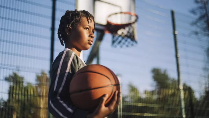 Young girl with dark skin tone holds basketball beneath a basketball hoop on a sunny day