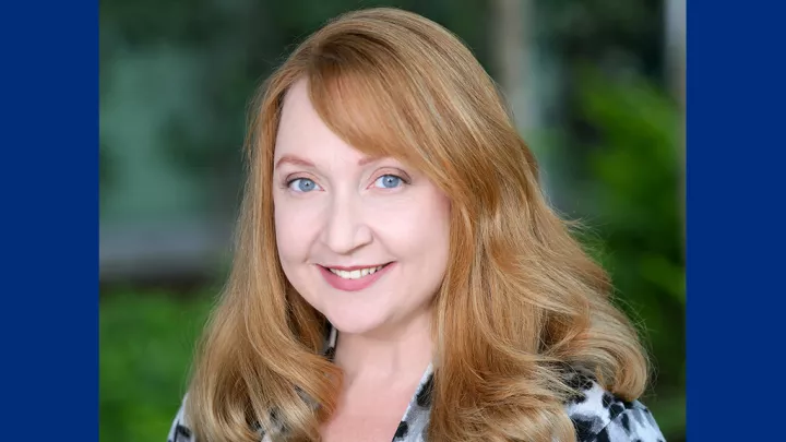 Headshot of a smiling woman with light skin tone and auburn hair wearing a black and white patterned jacket against a blurred outdoor background