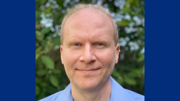 Headshot of a smiling man with light skin tone and light hair wearing a blue shirt against a blurred outdoor background