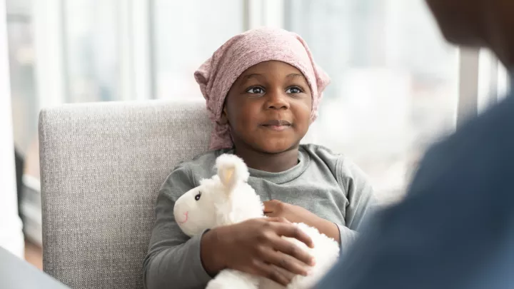 Young girl with dark skin and wearing a pink beanie clutches a stuffed lamb while sitting in a grey upholstered chair across from an adult in the near foreground