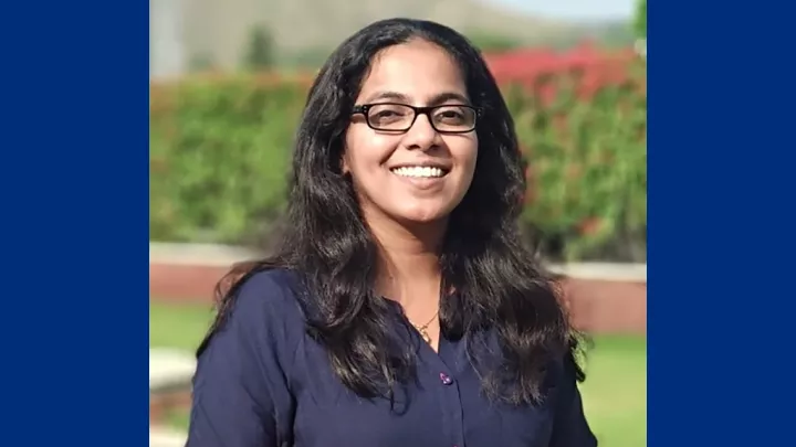 Headshot of a smiling woman with medium skin tone, long dark hair and glasses wearing a dark blue blouse against a blurred outdoor background