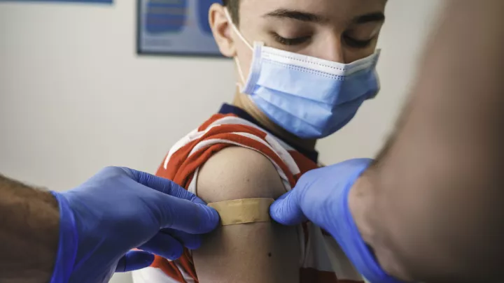 Health care worker with medium skin tone and wearing blue medical gloves places a bandaid on the right shoulder of a young boy with light skin tone and short brown hair wearing a surgical mask