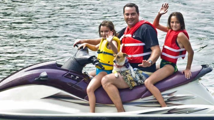 Father and two daughters with light skin and brown hair wearing life jackets sit on a Jet Ski in the water