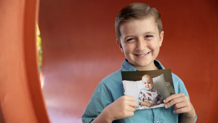 Young boy with light skin tone and brown hair wearing aqua colored short sleeved shirt smiles as he holds a picture of himself as a toddler undergoing treatment for cancer
