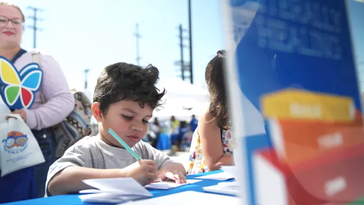 Young boy with medium skin tone and dark hair colors at an outdoor table as his mother watches over his shoulder