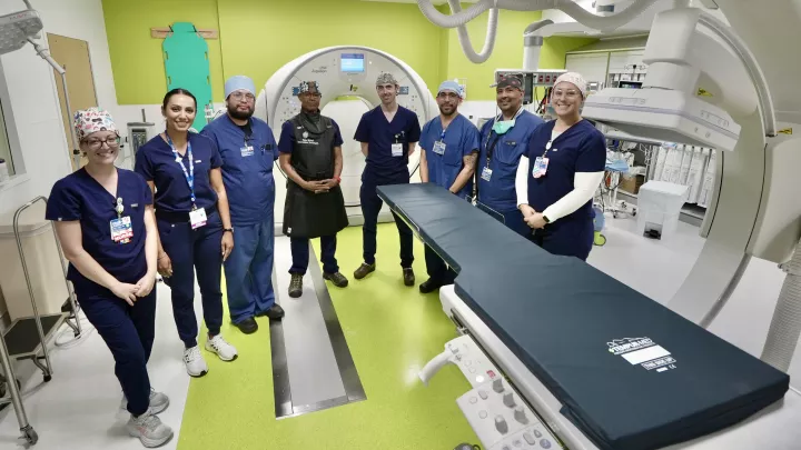 A group of smiling men and women wearing blue scrubs stand in a semi-circle around new CT-fluoroscopy machine