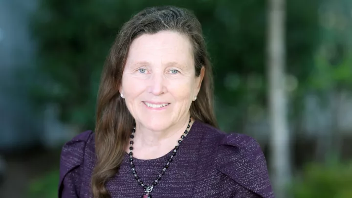 Headshot of a smiling woman with light skin and long brown hair wearing a beaded necklace and purple top against a blurred outdoor background