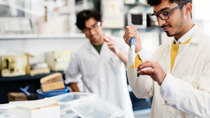 Student with medium skin tone and dark hair wearing black glasses and white lab coat learns how to use special laboratory equipment with professor watching in background