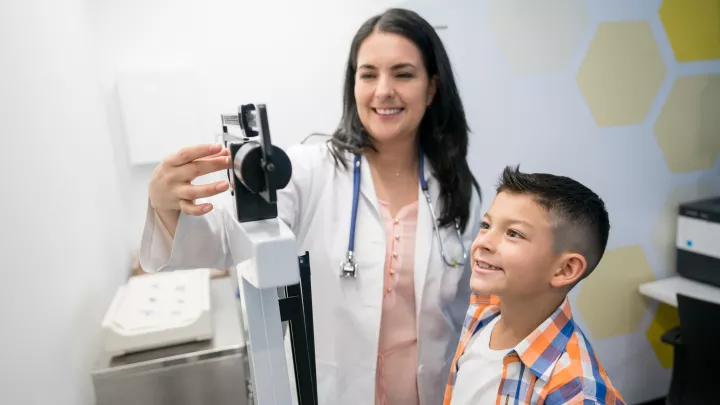 A light-skinned doctor in a white coat smiles while checking the weight of a boy with medium skin tone wearing a plaid shirt