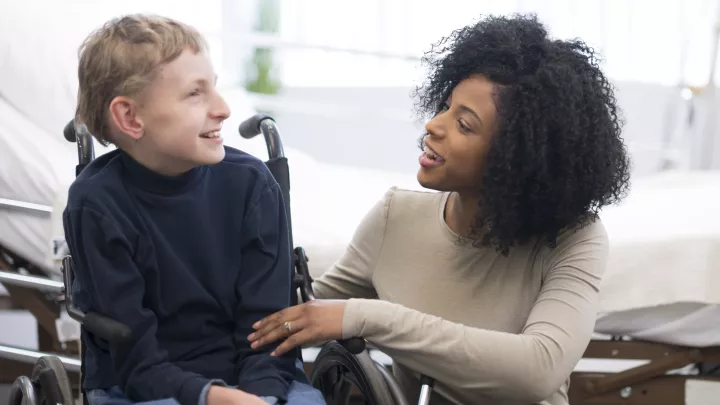 A female physical therapist with dark skin tone and dark hair works with a child with light skin tone and blonde hair seated in a wheelchair