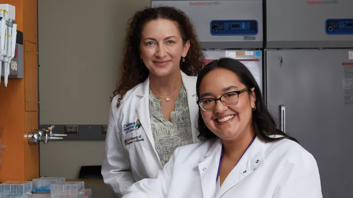 Two female researchers wearing white lab coats sit at a desk in a laboratory setting