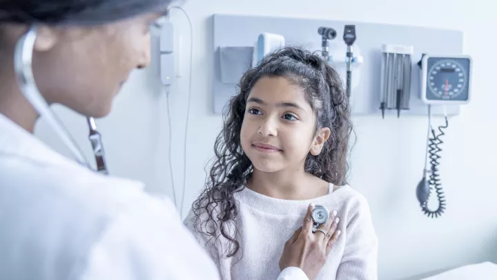 A female doctor with medium skin tone and dark hair listens to her young patient's heartbeat as she sits up on an exam table.