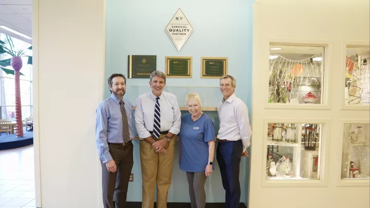 Three men and one woman in a hospital lobby smile at the camera in front of a plaque that reads ACS: Surgical Quality Partner. 