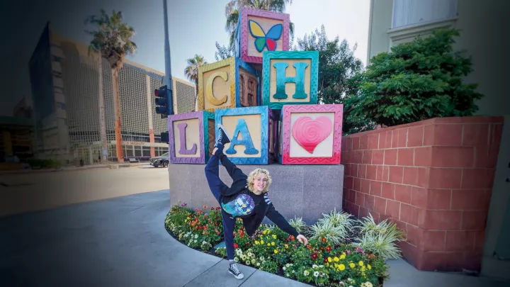 A smiling young woman with light skin tone and short blonde hair strikes a figure skating pose in front of Children's Hospital Los Angeles' iconic blocks statue