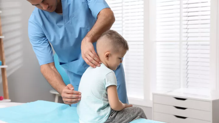 A doctor in blue scrubs checks a young boy’s back during an exam. The boy wears a light T-shirt and is sitting on an exam table