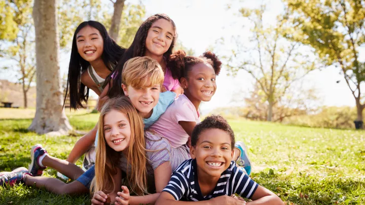 Six children with light, medium and dark skin tones and hair colors pile in a pyramid, smiling, on green grass in a park.