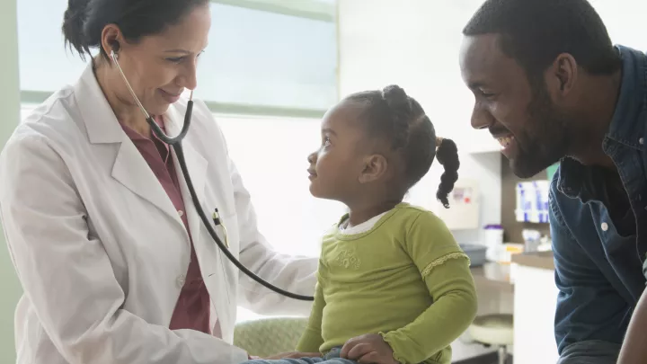 A female doctor with medium skin tone listens to the lungs of a little girl with dark skin tone and hair. The girl’s dad looks on, smiling.