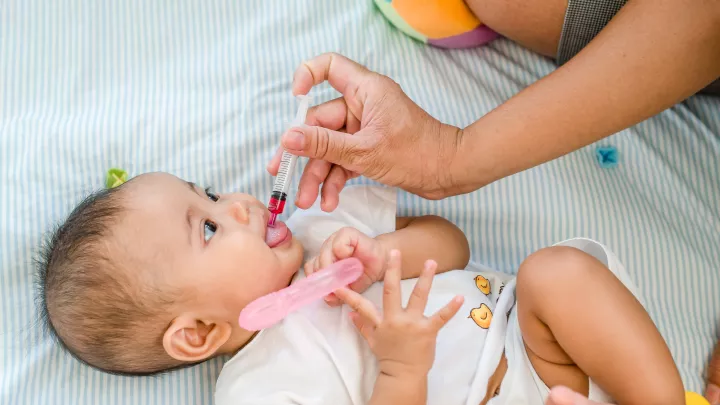 An adult hand syringes red liquid medicine into a baby’s mouth. The baby is looking at the caregiver and clutching a pink toy.