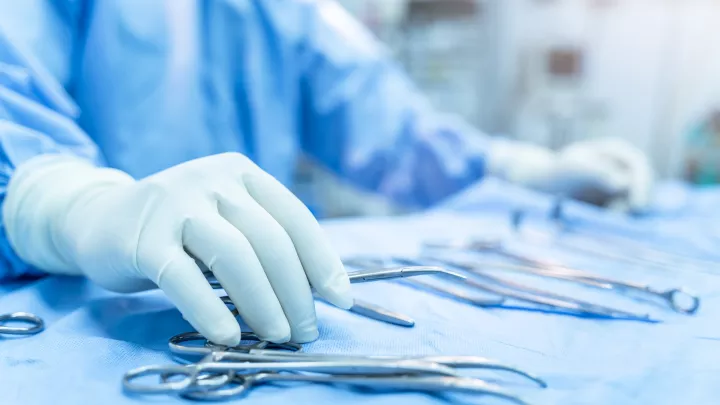 The gloved hand of a nurse wearing blue scrubs reaches for steel instruments laid out on a blue table in the operating room.