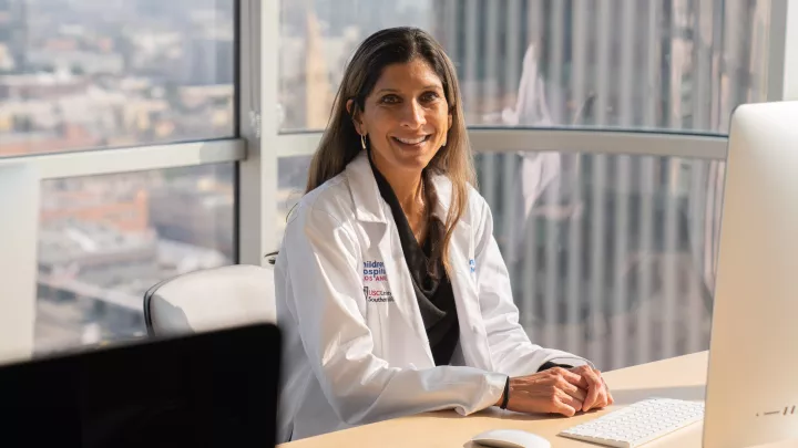 A woman with medium skin tone and long brown hair wearing a dark blouse under a white lab coat smiles as they sit at an office desk with Los Angeles visible through the large windows in the background
