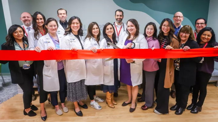 A group of 15 healthcare workers stand in a line behind a red ribbon as a female doctor with light skin tone and dark hair wearing a white lab coat gets ready to cut the ribbon with an oversized pair of scissors