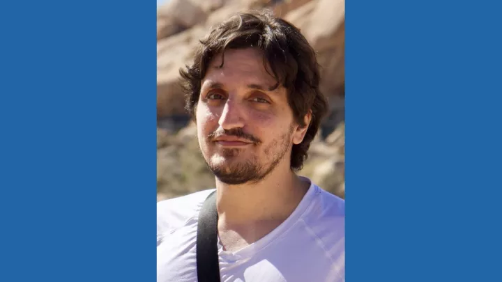 Headshot of a man with medium skin tone, brown hair and close-cropped brown beard wearing a white v-neck t-shirt against a blurred rocky, desert background