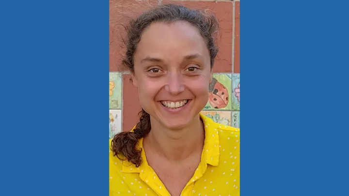 Outdoor headshot of a smiling woman with medium skin tone and brown hair pulled back into a ponytail wearing a bright yellow blouse against a red brick wall adorned with colorful tiles of children's drawings