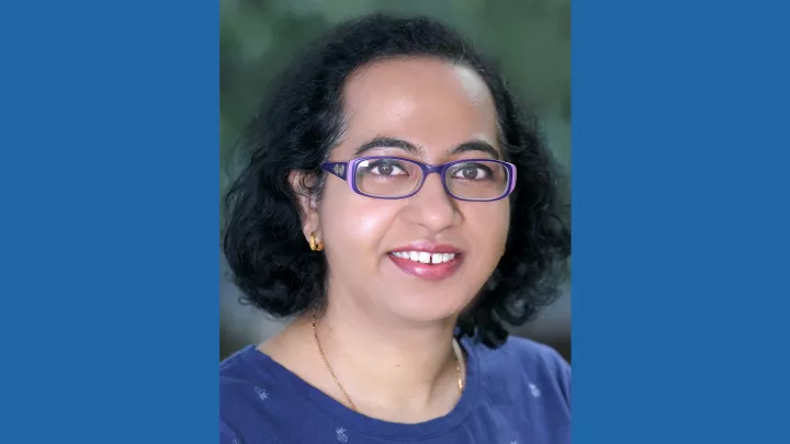 Headshot of a smiling woman with medium skin tone and dark hair wearing purple rectangular eyeglasses, gold earring and necklace and a top top against a blurred outdoor background