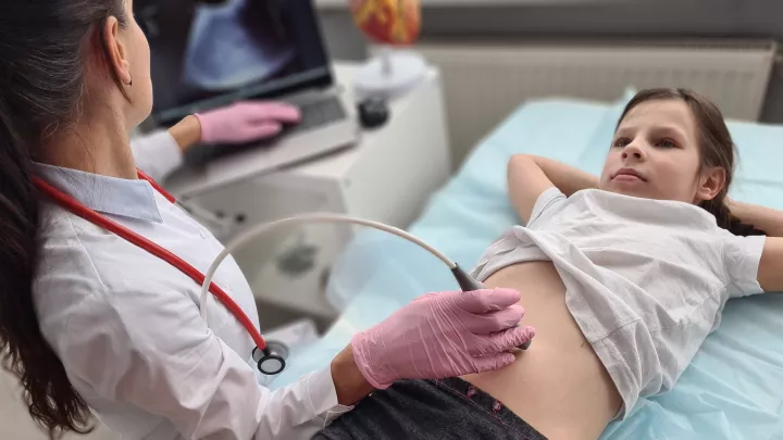 A woman in a lab coat and latex gloves uses an ultrasound transducer on the stomach area of a light skin-toned girl laying on an exam table.