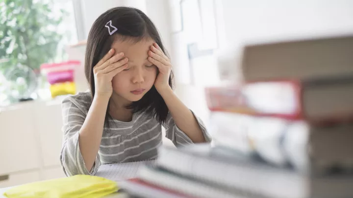 Young girl with medium skin tone and dark hair rubs her forehead as she sits behind stack of textbooks