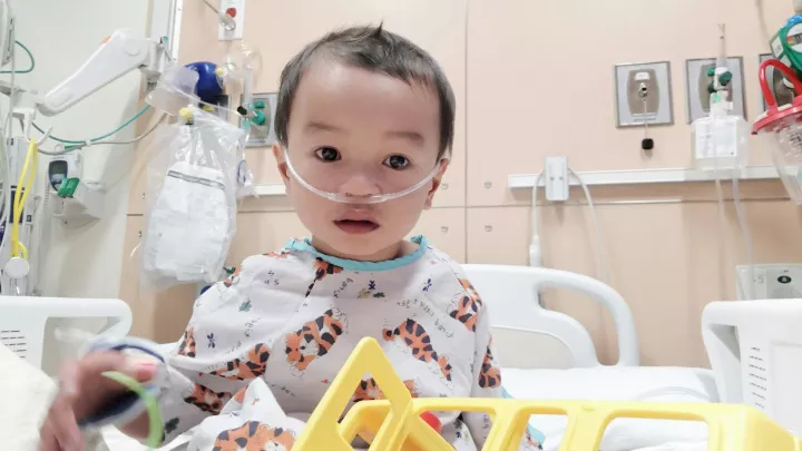 A toddler with medium-light skin tone sits in a hospital bead, wearing hospital pajamas with a nasal cannula in his nose.