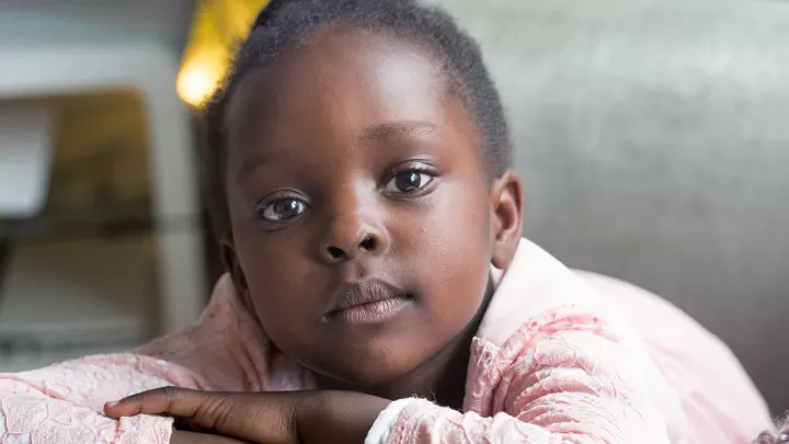 Young girl with dark skin tone and short dark hair wearing a pink sweater sitting in a grey chair looking at camera