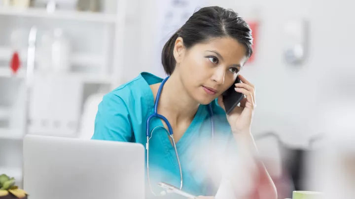 Medium-light skin-toned woman in scrubs with a stethoscope around her neck talks on a cell phone. She appears irritated.