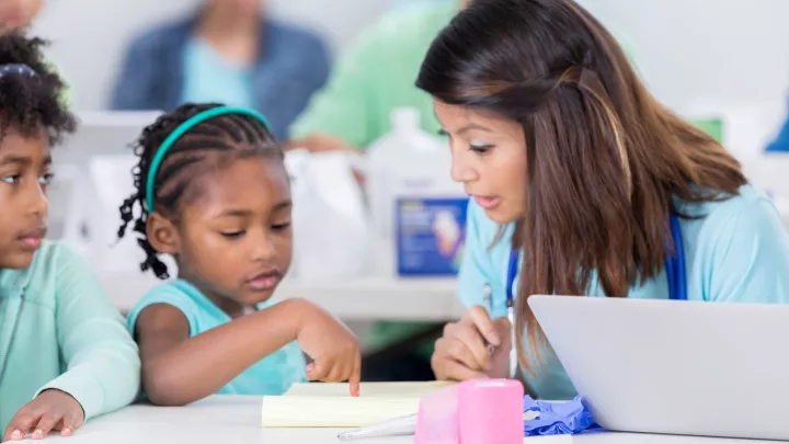 Two young girls with medium-dark skin tone sit at a table. One of them points at a pad of paper while a medium skin-toned woman looks at where she is pointing