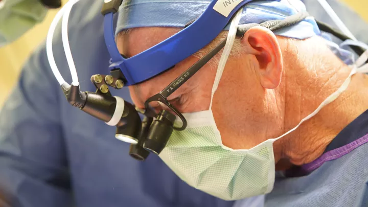 Close-up image of Vaughn Starnes, MD as he performs procedure wearing a surgical mask, cap, gown, surgical loupes and a headlight