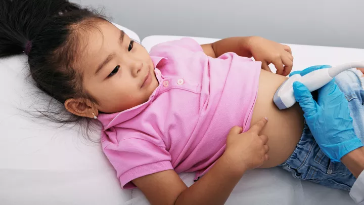 Young girl with medium skin tone and dark hair lies on her back and pulls up her pink shirt so a gloved health care provider can use ultrasound to scan her lower abdomen