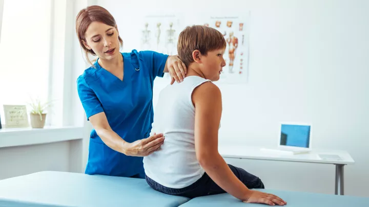 A doctor in blue scrubs checks a teen boy’s spine. The boy has light skin and hair and is sitting on a blue exam table.