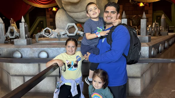 Father with medium skin tone and dark hair smiles as he holds son with medium skin tone and short brown hair and poses with two young daughters in front of decorative altar