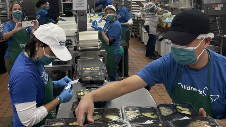 People in caps, surgical masks and blue shirts prepare and wrap food in an industrial kitchen