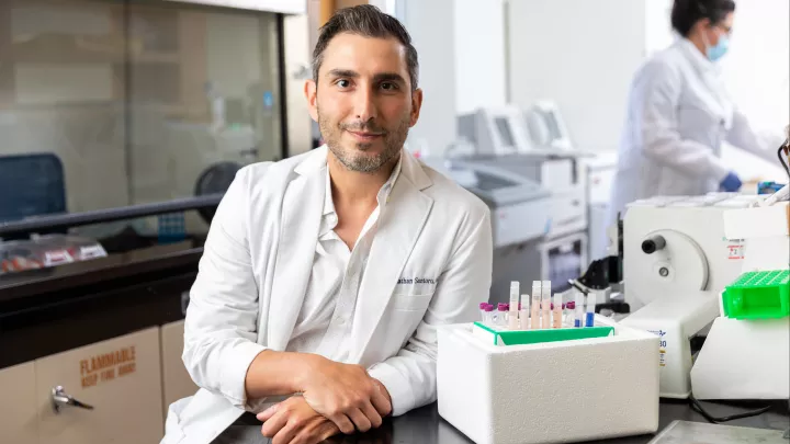 A man in a lab coat leans on a research bench, smiling at the camera