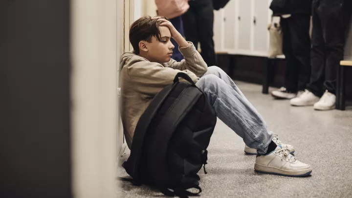 Side view of a medium skin-toned teenage boy sitting against his locker next to his backpack in a crowded school hallway, looking exasperated