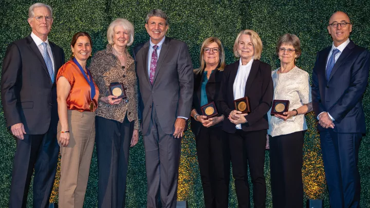 Five women and three men in business attire stand in a row against a green background